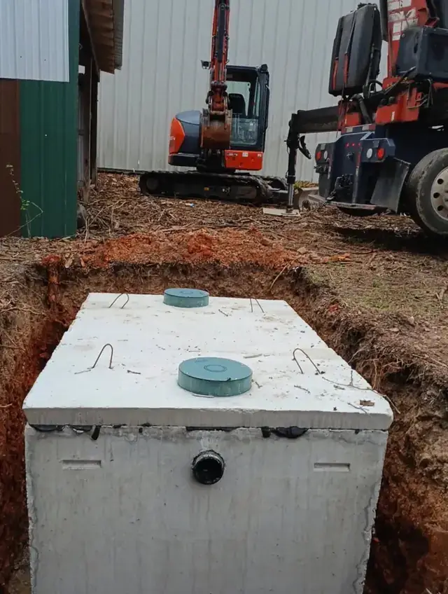 A freshly set concrete septic tank with green risers in a red clay excavation, an orange mini excavator behind it