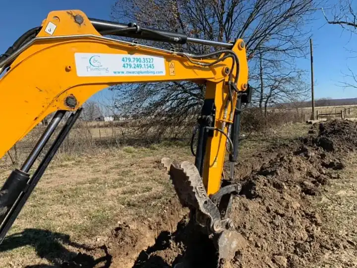 A yellow mini excavator arm with the C&S Plumbing decal digging a trench across a pasture