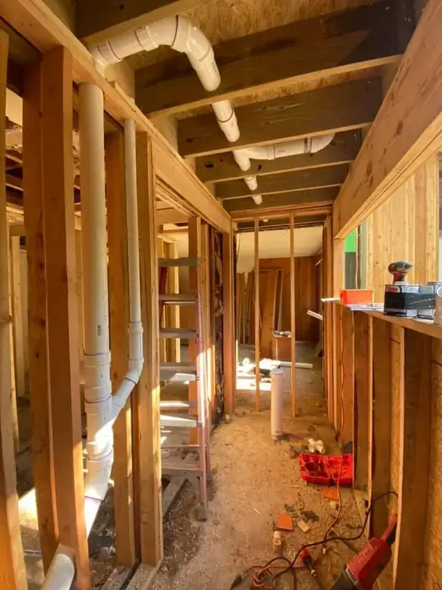 White PVC drain stack and vents running down a framed hallway on a remodel rough-in with a red tool case on the subfloor