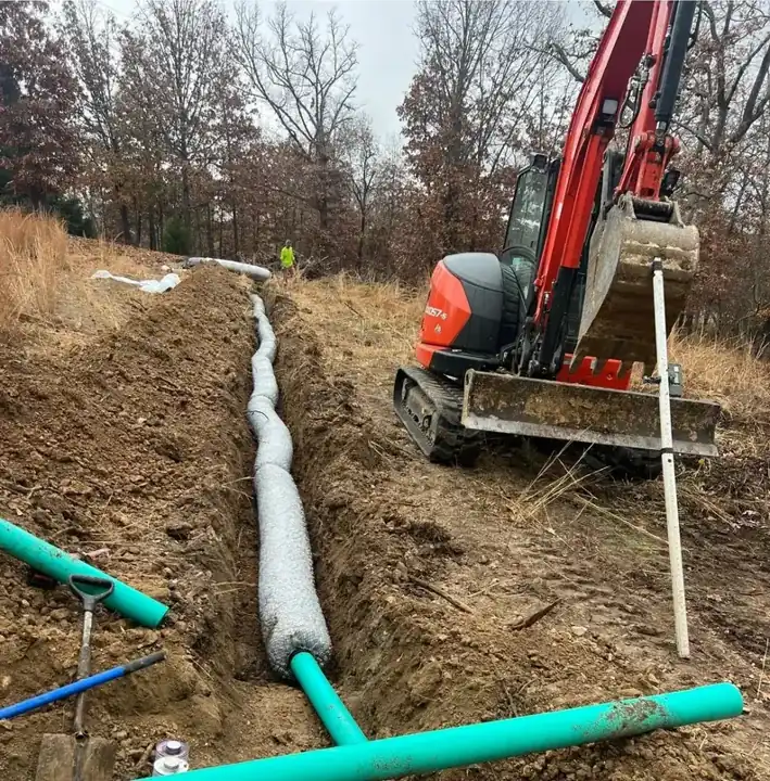 Drain field line wrapped in filter fabric laid in a fresh trench with a green PVC header and a red mini excavator