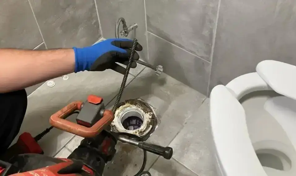 Gloved hand feeding a drain auger into an open toilet flange on a tiled bathroom floor
