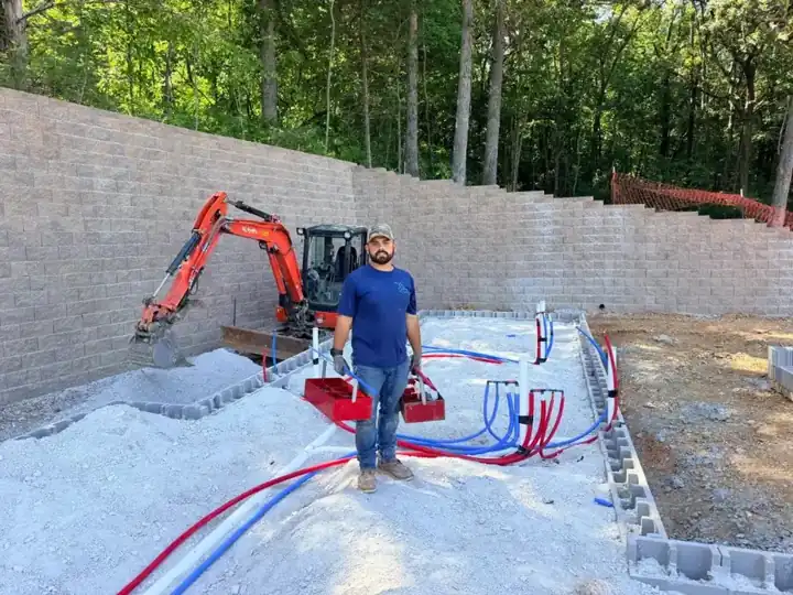 A C&S plumber standing on a gravel slab pad among red and blue water lines, a mini excavator and a block retaining wall behind him