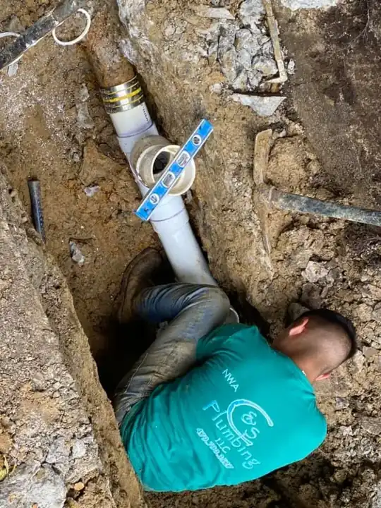 A technician in an NWA C&S Plumbing shirt standing in a trench leveling a PVC sewer tie-in
